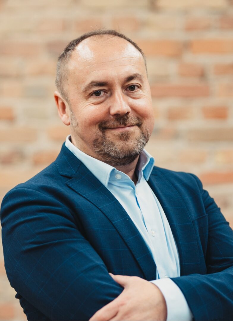 Uśmiechnięty mężczyzna z brodą. Ma założone ramiona. Ubrany w garnitur i błękitną koszulę.